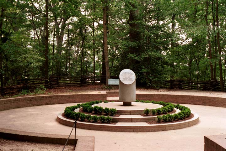 A circular brick patio, about 40 feet in diameter, with a foot-high brick wall surrounding and a cylindrical pillar in the center, about three feet high with a marble plaque set at a 70 degree angle. The pillar is set in the center of a three-tiered circular stepped area, with brick walls with small shrubs on the first and second tiers. The back of the area is wooded with a split-rail fence at the edge. (103K)